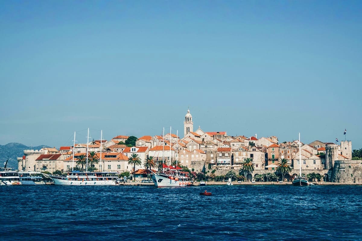 Historic coastal town of Dubrovnik viewed from the sea with sailboats, terracotta roofs, and bell tower against clear blue sk