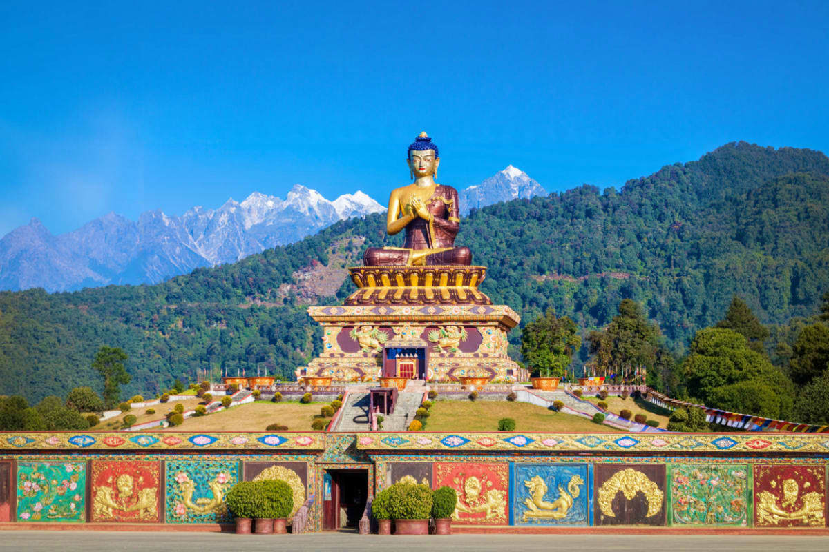 Golden Buddha statue seated on ornate pedestal with snow-capped Himalayan mountains and forested landscape in background unde