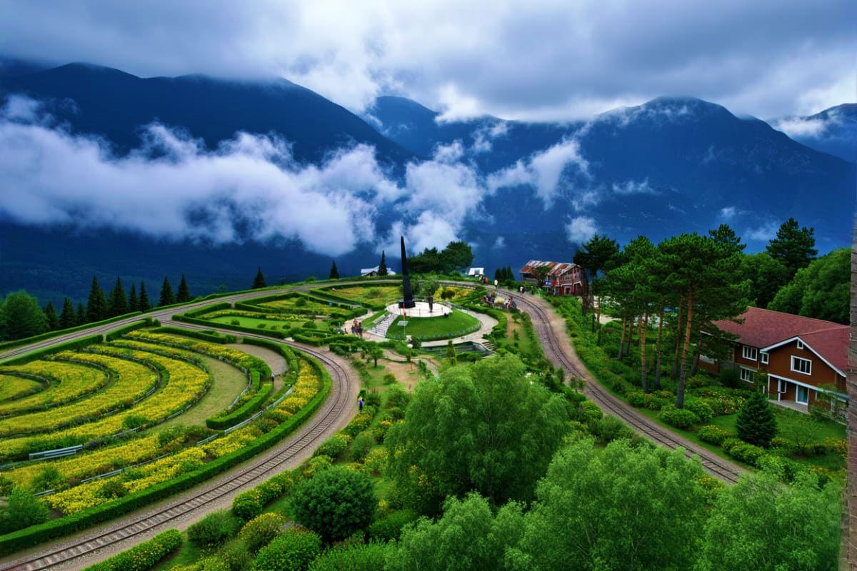Aerial view of a scenic mountain landscape with spiral garden maze, train tracks, traditional houses, and mist-covered Himala