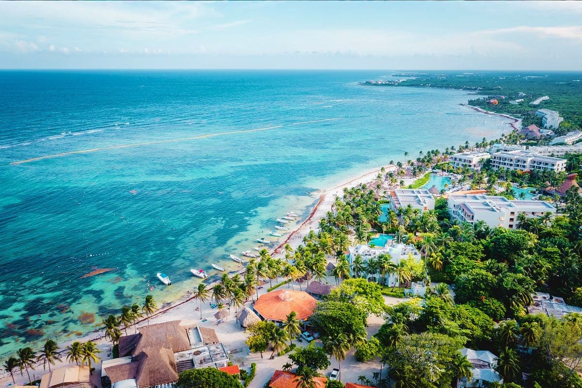Aerial view of a tropical beachfront resort with white buildings, pools, palm trees, turquoise ocean, and sandy beach with bo