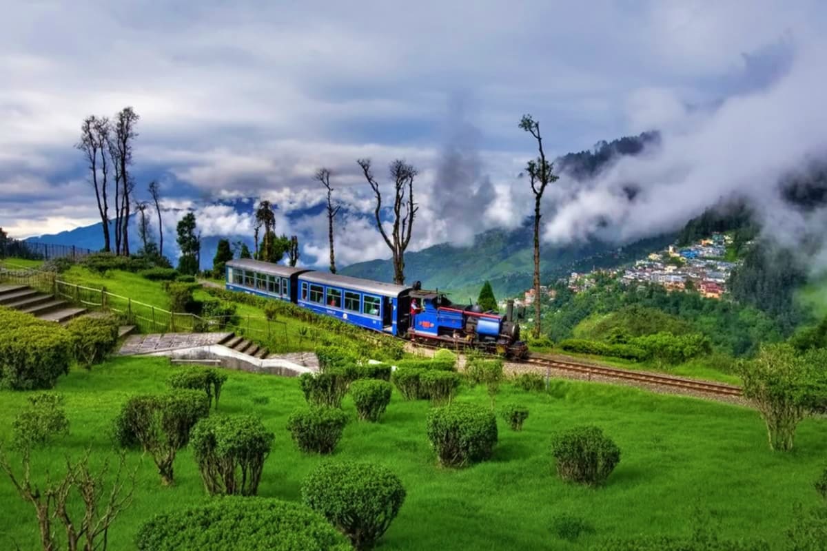 Toy train with blue passenger car traveling through green hillside landscape with misty mountains and Darjeeling town in back