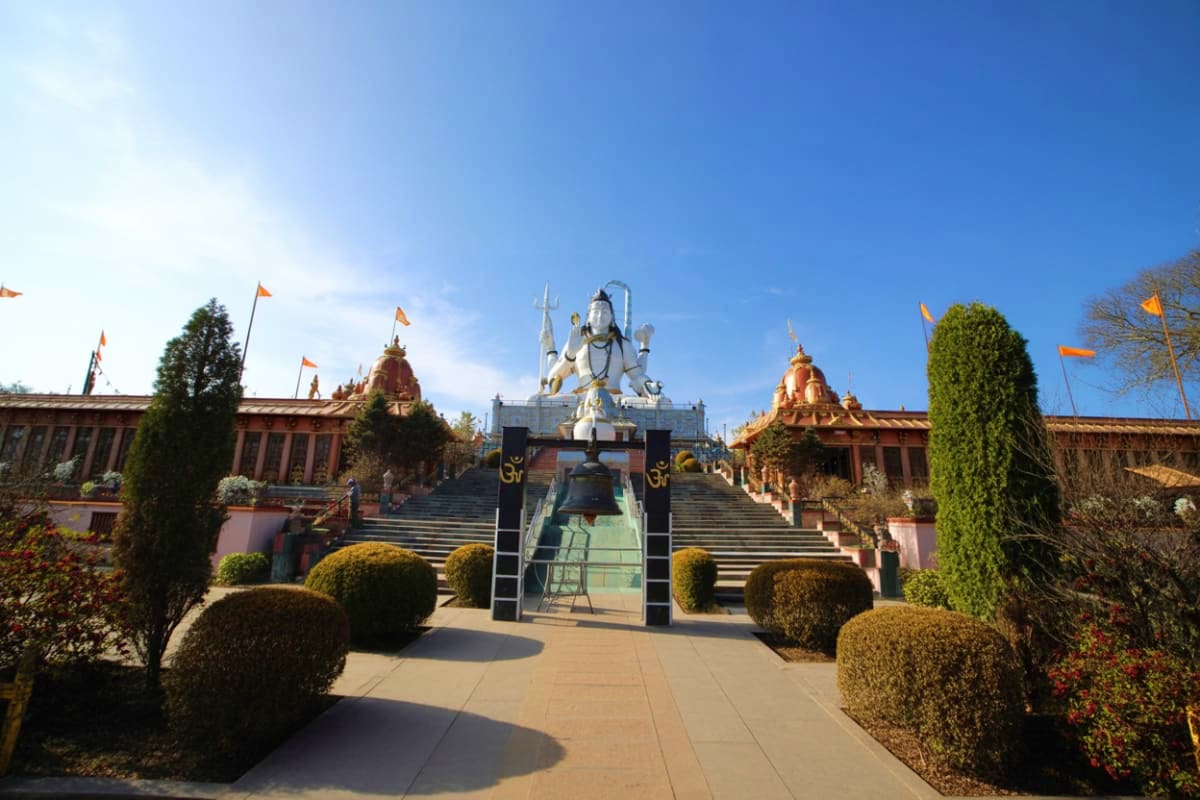 Grand white temple with golden spires at center of ornate religious complex with red buildings and Indian flags, surrounded b