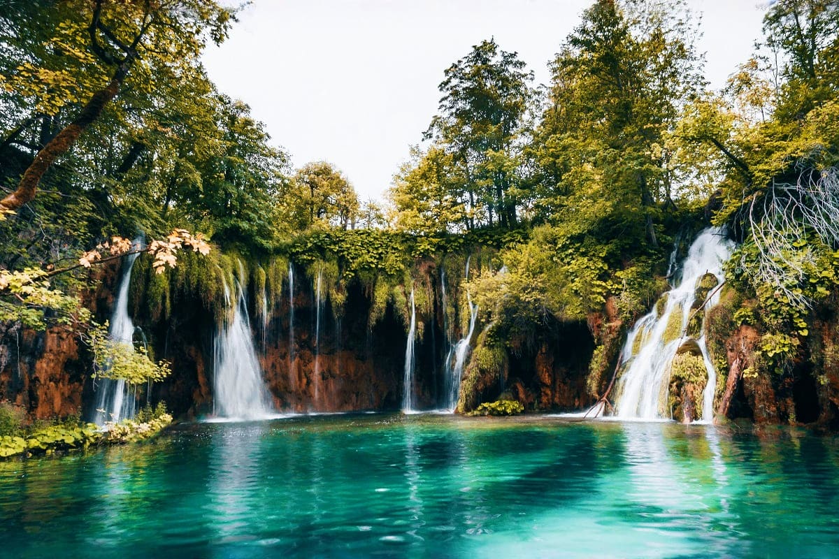 Multiple waterfalls cascading into a turquoise lake surrounded by lush green forest and moss-covered cliffs in Plitvice Lakes