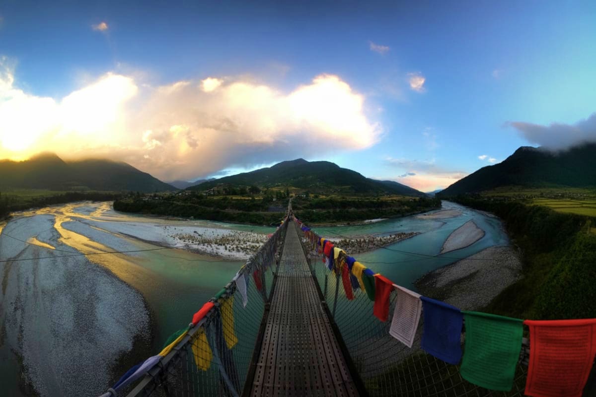 Suspension bridge with prayer flags spanning turquoise river valley surrounded by mountains in Bhutan