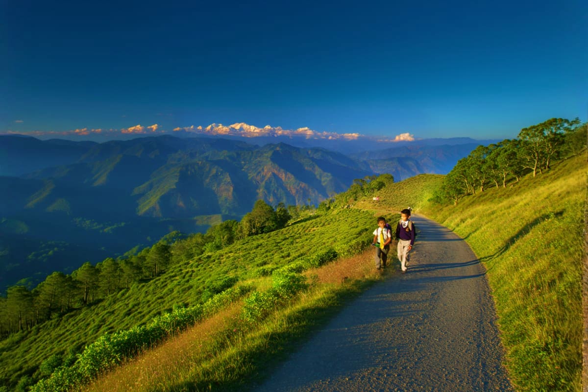 Two hikers walking on a mountain road with lush green valleys and distant Himalayan peaks under blue sky in Bhutan