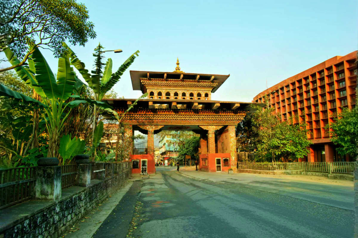 Traditional Bhutanese gateway with ornate golden roof and red buildings, featuring tropical plants and modern architecture in