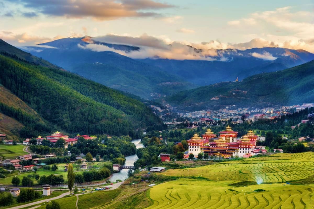 Scenic valley view of Bhutanese monastery with terraced rice fields, river, forested mountains and snow-capped peaks at sunse