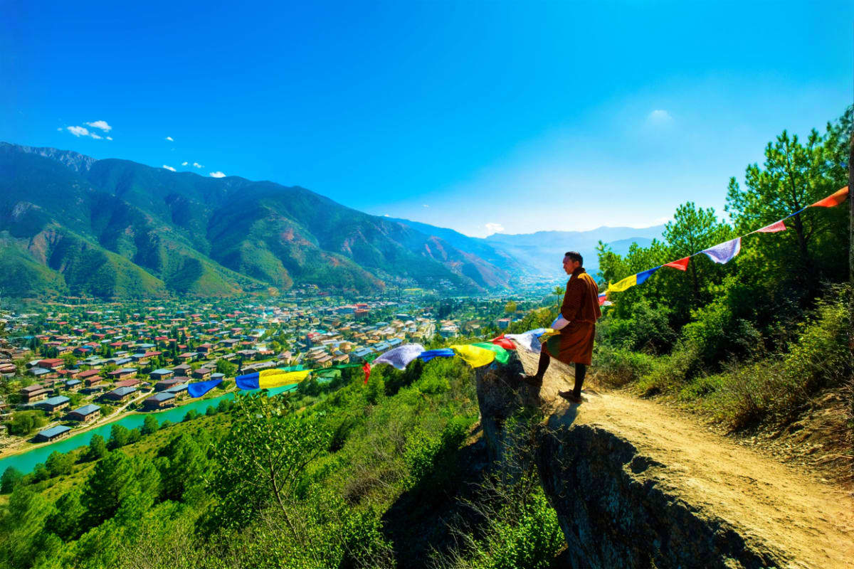 Person holding colorful prayer flags on mountain overlook above Bhutanese valley with turquoise river and town below