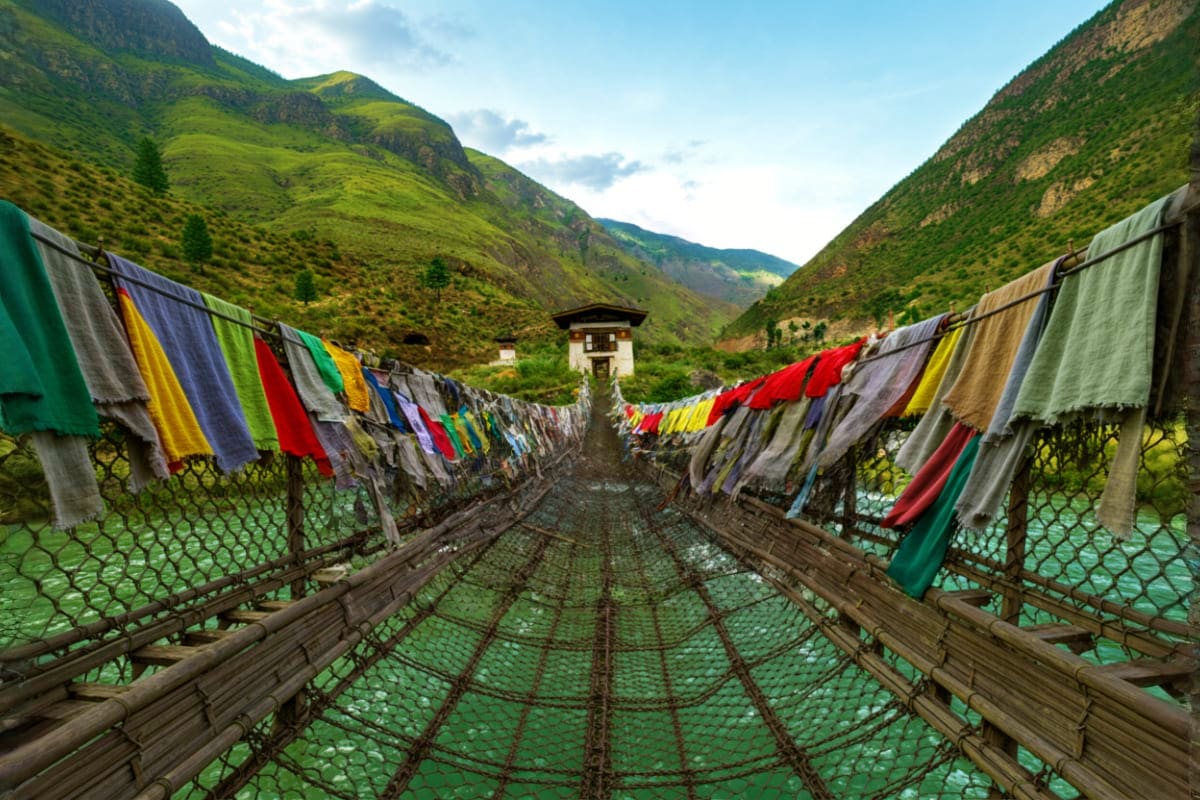 Colorful prayer flags hanging on wooden suspension bridge over emerald river with green mountains and traditional Bhutanese t