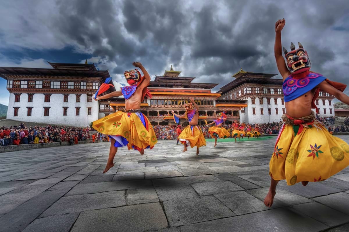 Performers in traditional Bhutanese costumes with colorful masks and yellow skirts dancing at Tashichho Dzong during a festiv