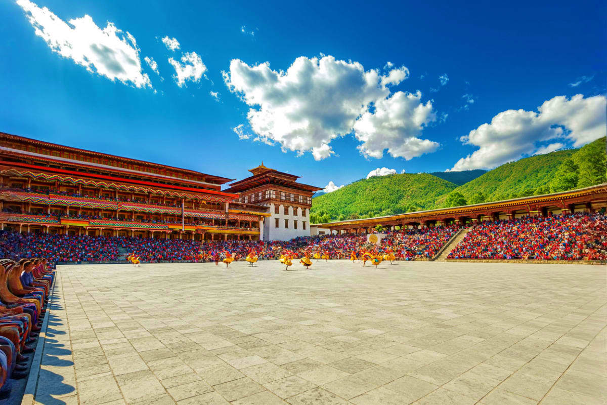 Taksang Dzong festival ground with ornate red and gold monastery architecture, performers in yellow robes, spectators in stan