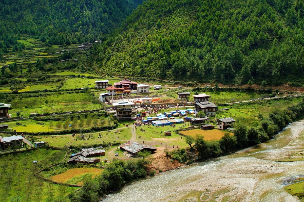 Aerial view of a Bhutanese monastery complex nestled in a lush green valley surrounded by dense conifer forests and a glacial