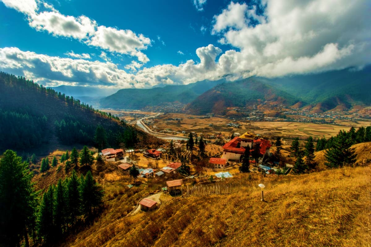 Phobjikha Valley landscape with traditional red-roofed farmhouses, river, forested mountains, and golden fields under blue sk