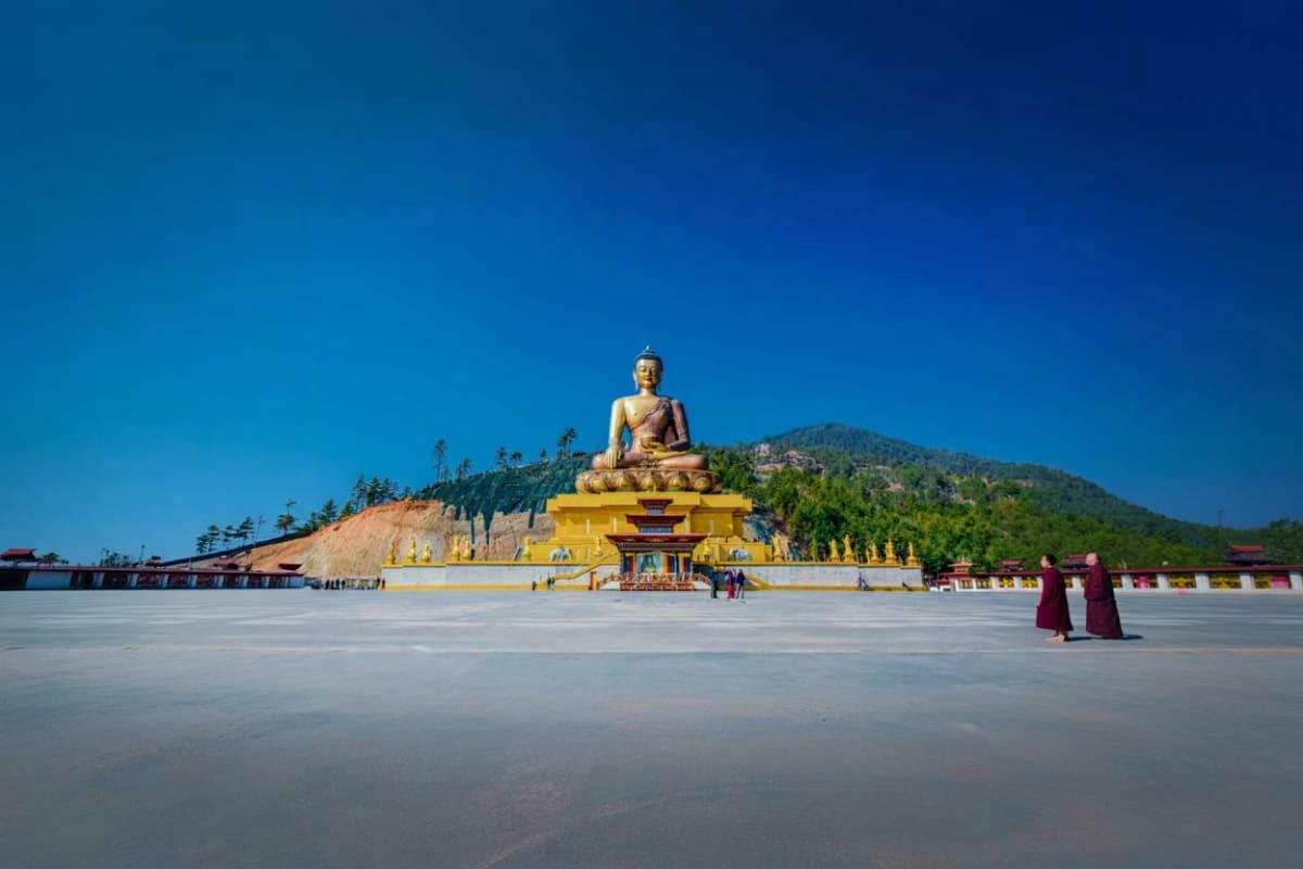 Giant golden Buddha statue seated on yellow platform with mountain backdrop and monks walking in courtyard