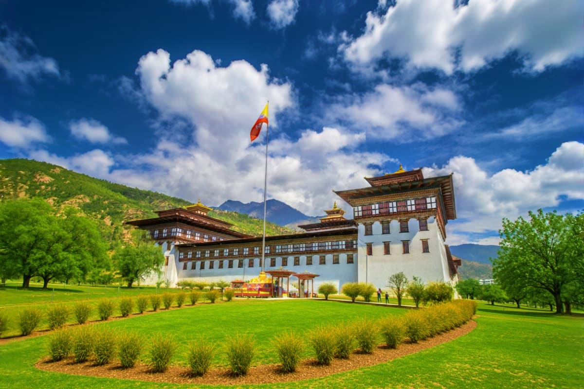 Rinpung Dzong fortress in Bhutan with Bhutanese flag, green meadow, and mountain backdrop under blue sky with clouds