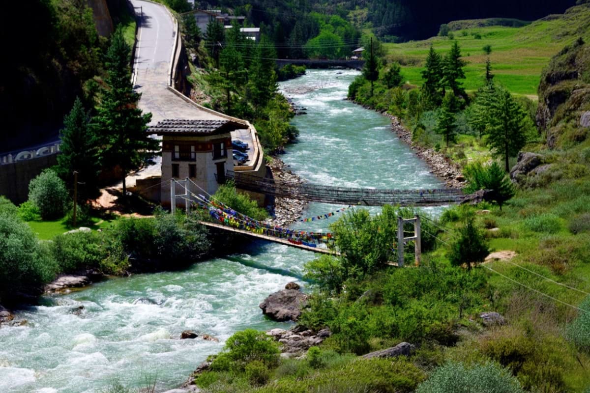 Turquoise river flowing through forested valley with suspension bridge, traditional building, and winding mountain road in Bh