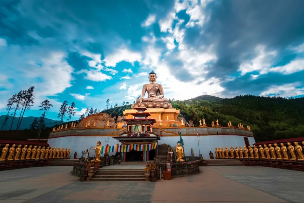 Large golden Buddha statue seated on orange temple structure surrounded by golden statues and prayer flags in Bhutanese mount