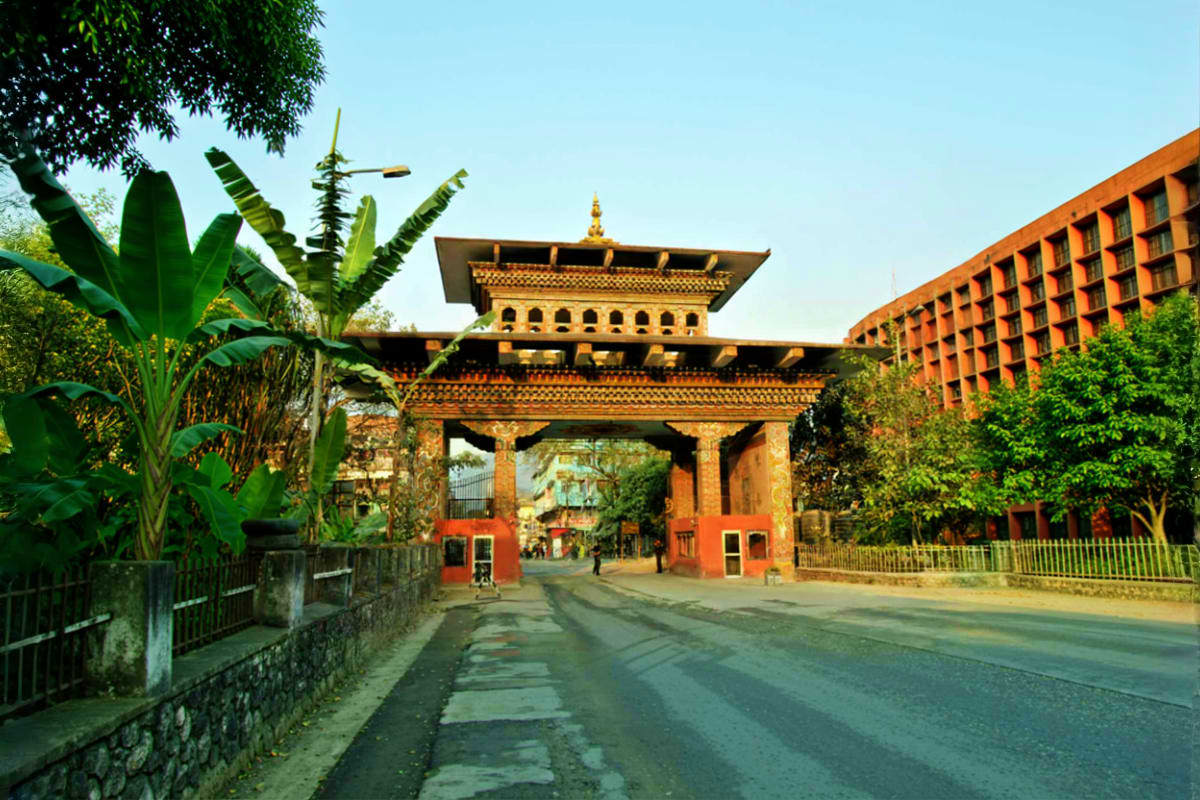 Traditional Bhutanese temple gate with golden roof and ornate architecture, flanked by tropical plants and modern buildings u