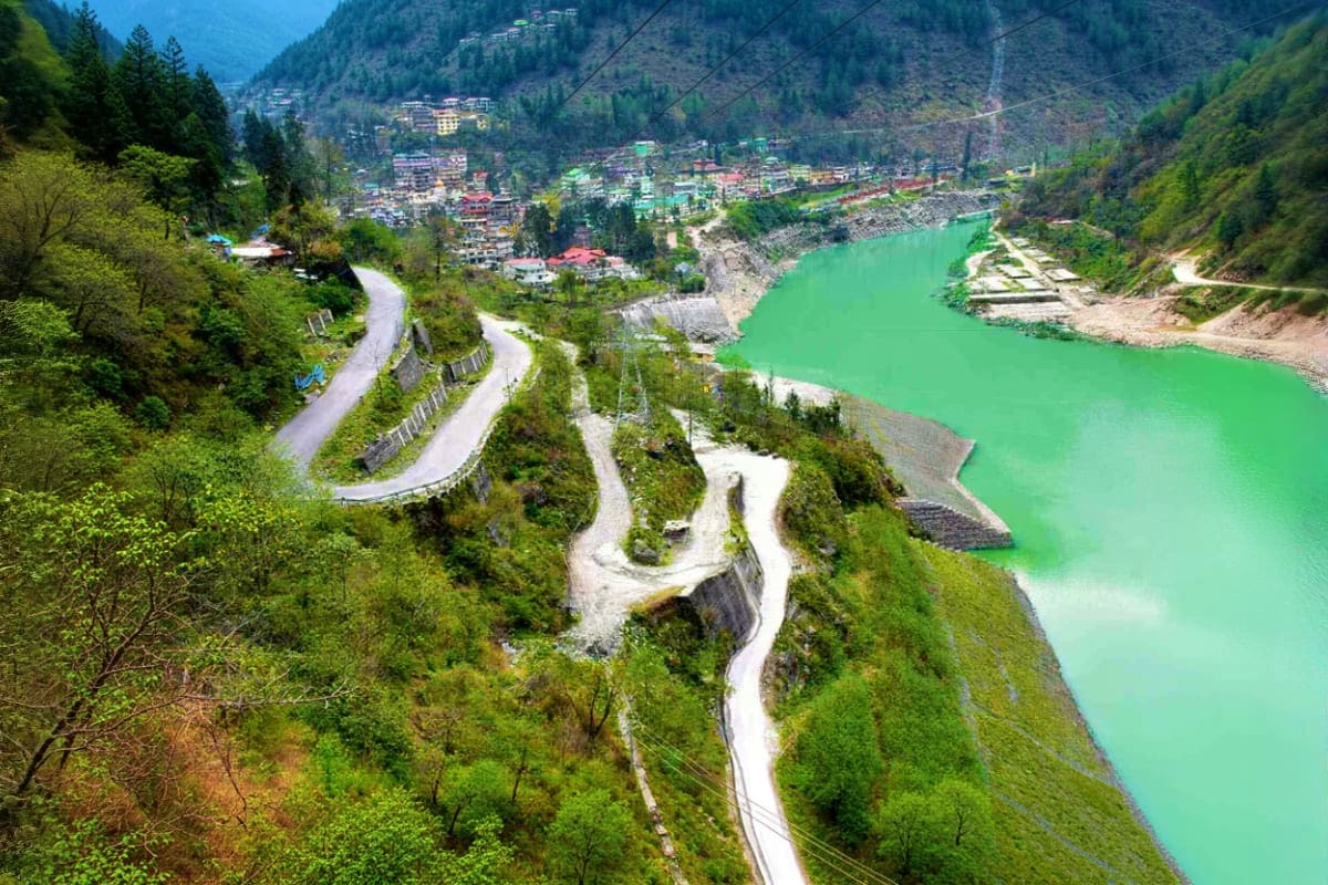 Aerial view of turquoise river winding through forested mountain valley with switchback road and town nestled in Himalayan la