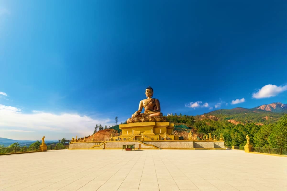 Giant golden Buddha statue seated on yellow platform overlooking forested mountains under blue sky in Bhutan