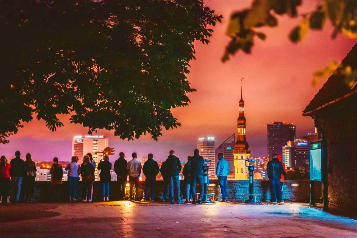 Group of tourists viewing Tallinn skyline at sunset with illuminated church spire and historic buildings reflected in water