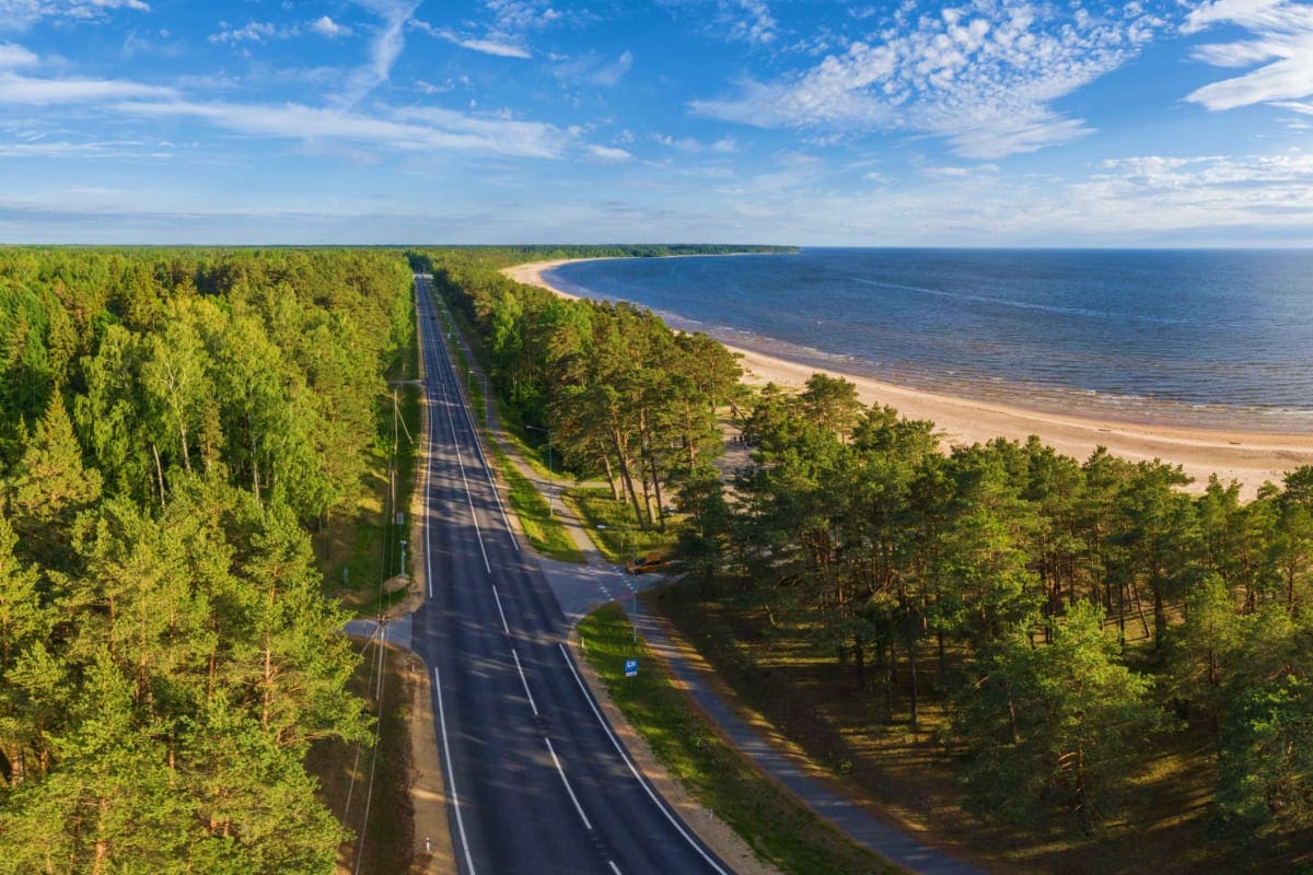 Aerial view of a straight highway cutting through green forest toward a sandy beach and blue sea coast in Estonia