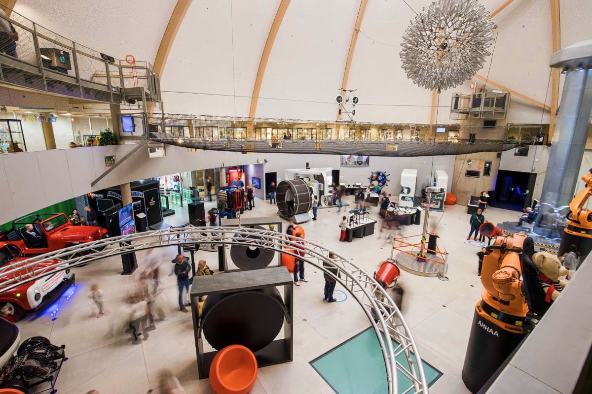 Modern interactive science museum exhibition hall with visitors exploring mechanical engineering displays, red vehicles, and 