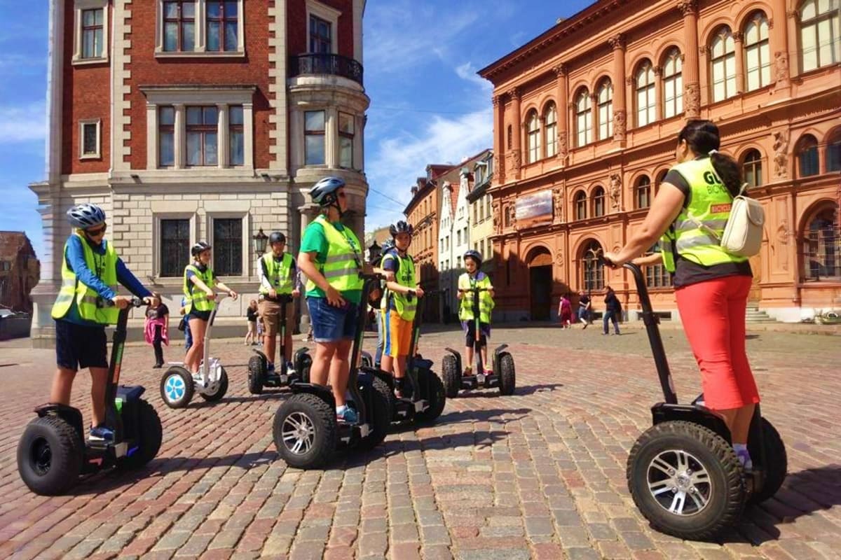 Group of tourists on Segways wearing yellow safety vests in historic town square with red brick buildings under blue sky