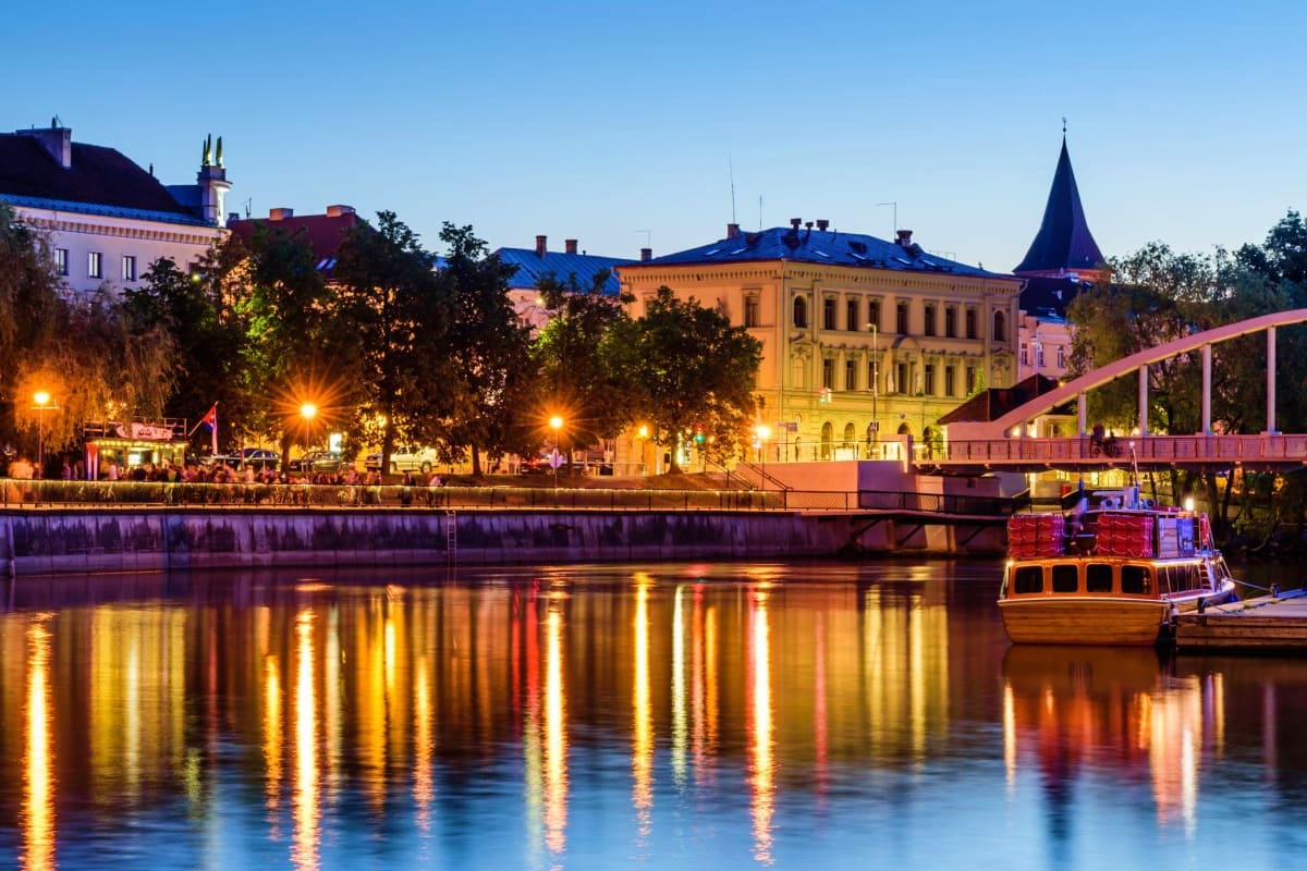 Evening waterfront view of Tartu with illuminated historic buildings, river reflections, and traditional wooden tour boat doc