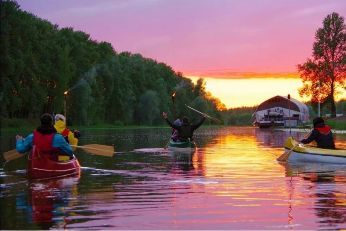Group of kayakers paddling on a calm river at sunset with tree-lined banks and a riverboat in the background during golden ho