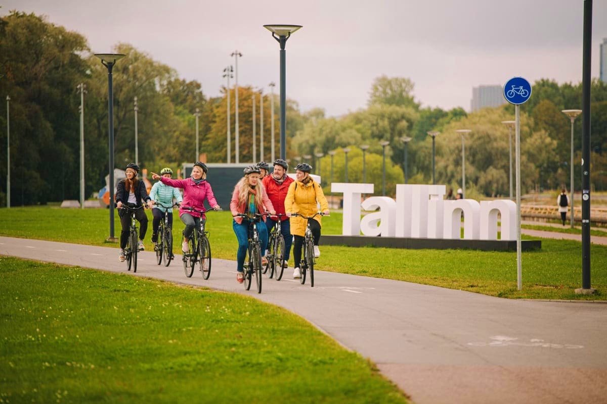 Group of cyclists riding on a paved path past the Tallinn sign in a park with green grass and trees
