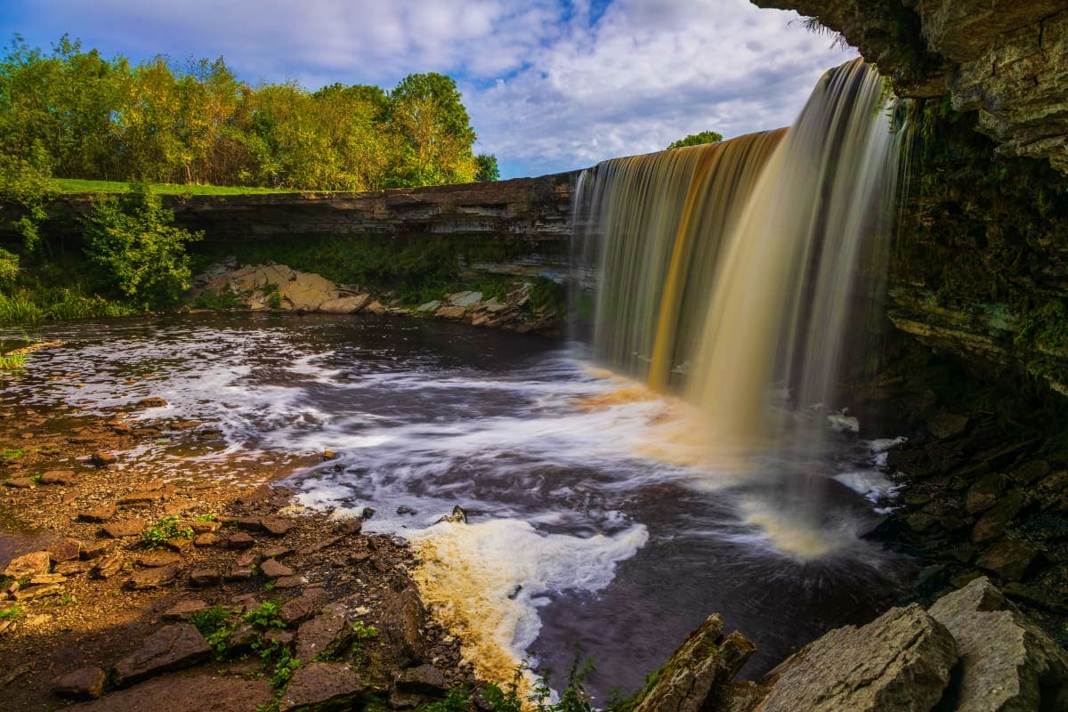 Waterfall cascading into rocky river pool surrounded by green trees and stone cliffs under blue sky with clouds
