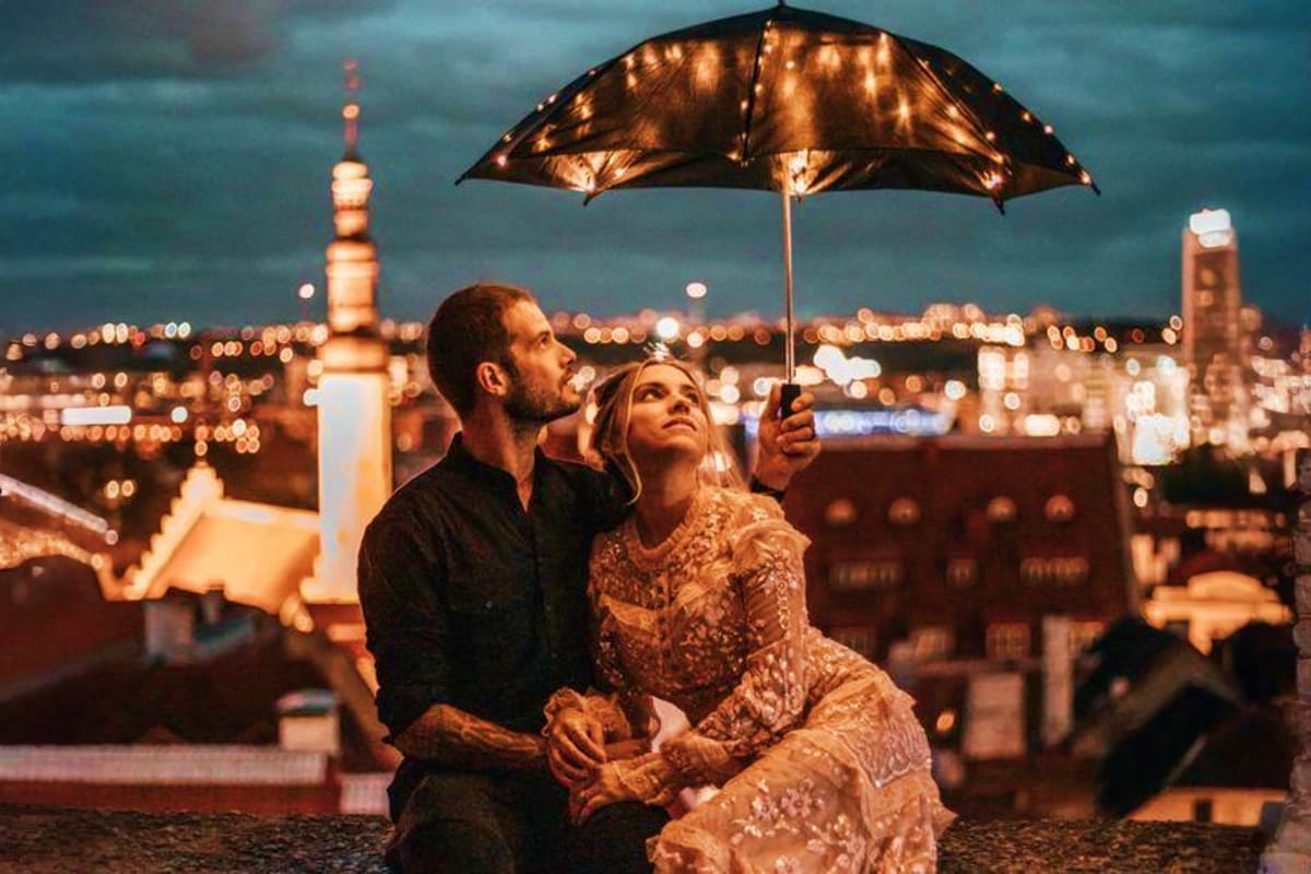 Couple holding illuminated umbrella on rooftop overlooking Tallinn cityscape at dusk with church spire and buildings lit up