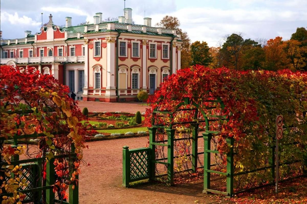 Red and cream classical palace with green roof surrounded by autumn foliage and green garden arbors with red ivy