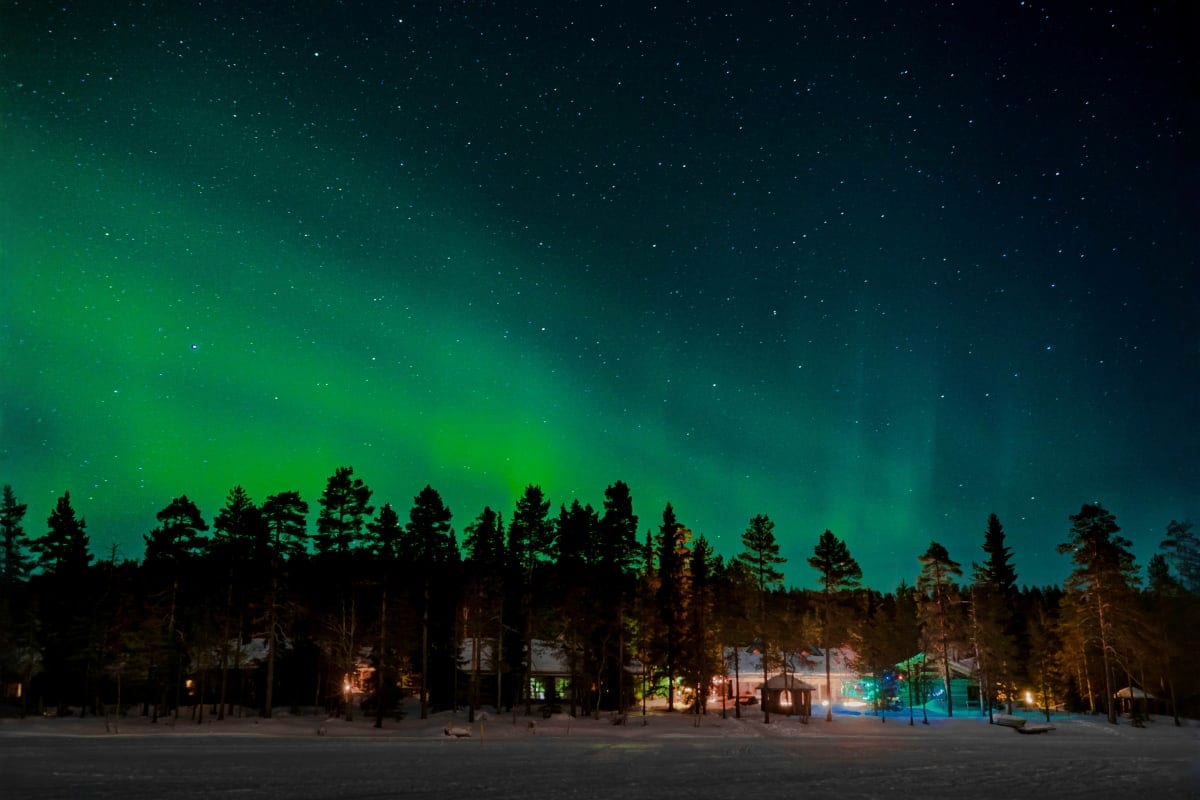 Northern lights dancing above snowy forest landscape with illuminated cabins and evergreen trees at night in Finnish Lapland