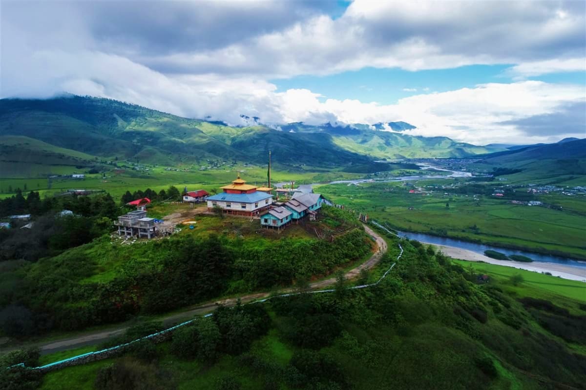 Aerial view of a Buddhist temple with golden spire on a hilltop overlooking green valleys, mountains, and a river in the Hima