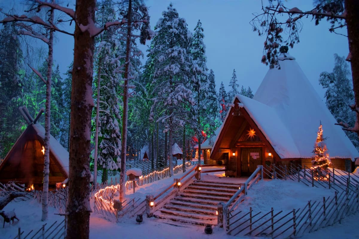 Snow-covered forest village at dusk with illuminated log cabins, wooden railings, decorated Christmas tree, and snow-laden ev