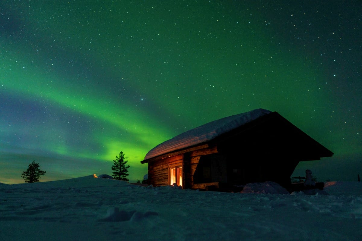 Wooden cabin glowing with warm light under vibrant green northern lights and starry sky in snowy Finnish landscape