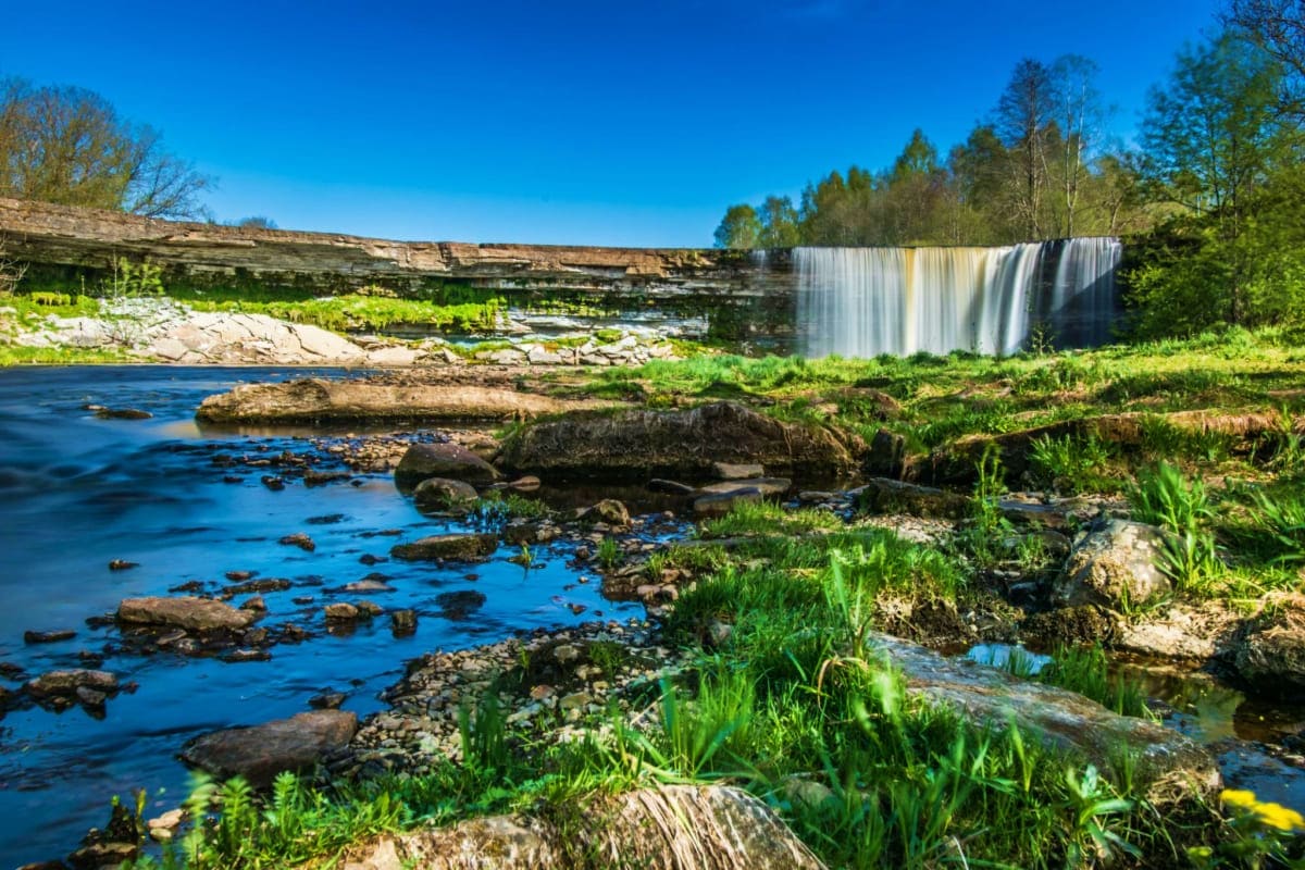 Scenic waterfall cascading over layered rock cliffs into a blue river with boulders and green vegetation in an Estonian fores