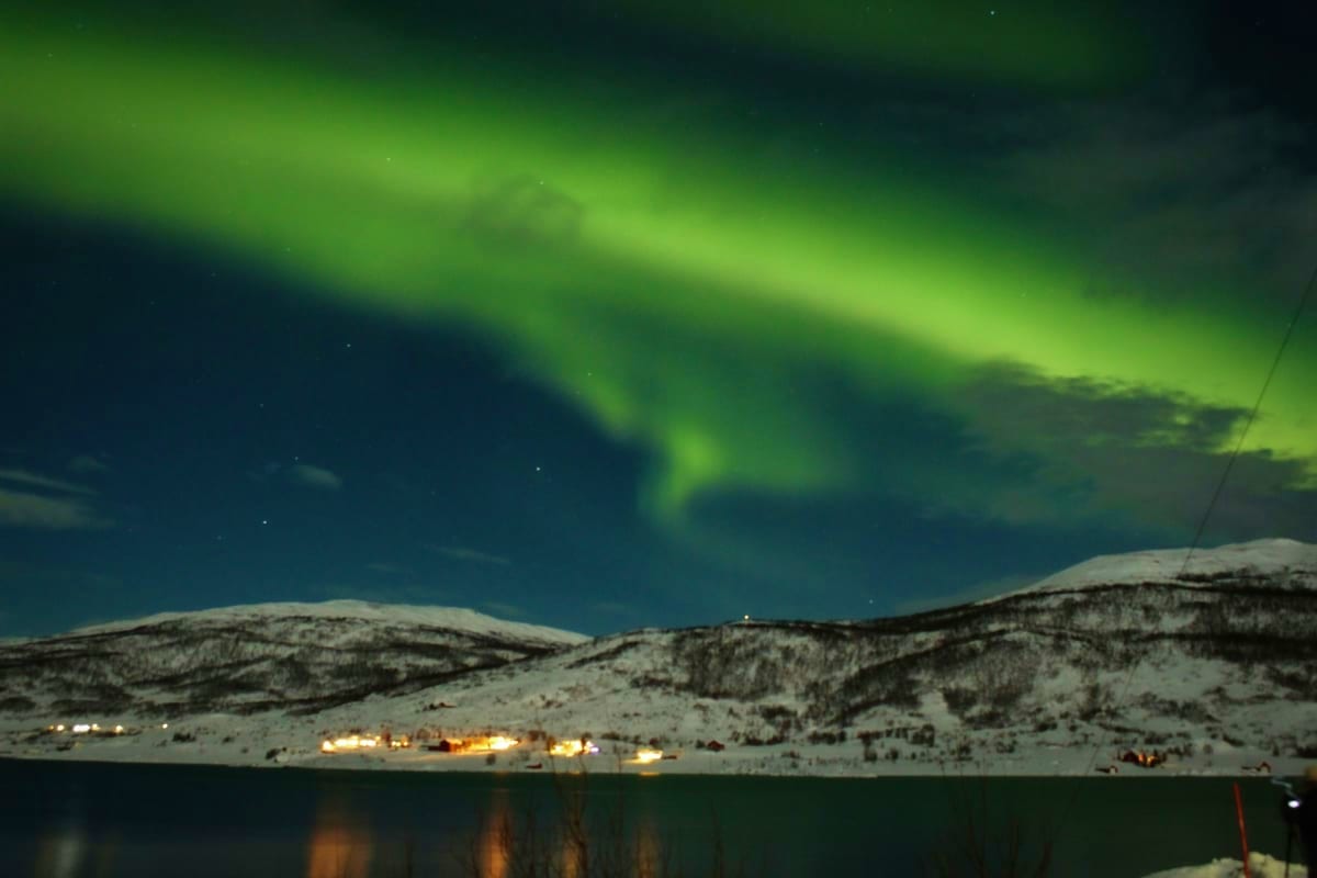 Northern lights dancing in green waves above snow-covered mountains and a small village with glowing lights by a frozen lake 