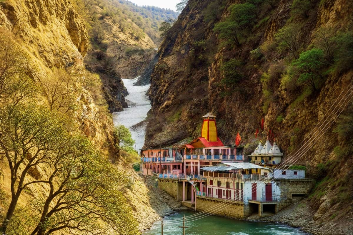 Colorful temple complex nestled in deep rocky canyon beside flowing river with green forest hills above, Char Dham pilgrimage