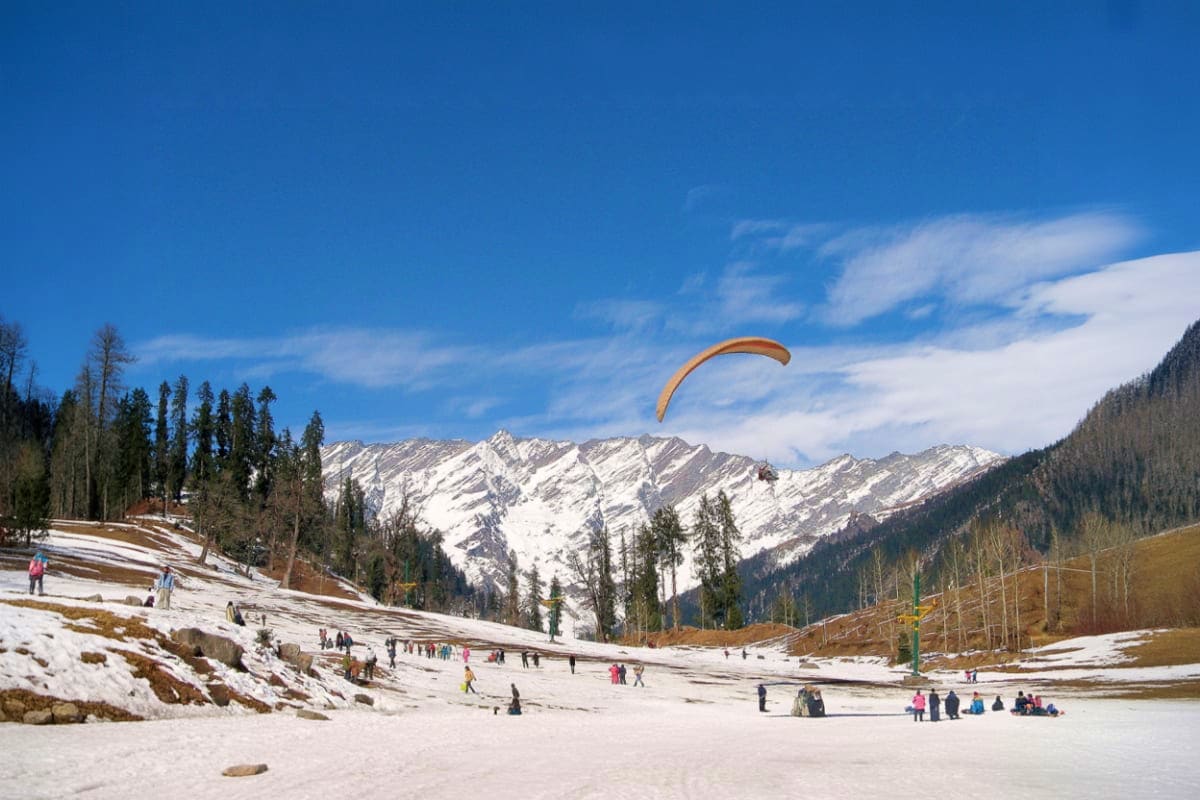 Paraglider flying over snowy Himachal Pradesh meadow with snow-capped mountain peaks and pine forests in background, tourists