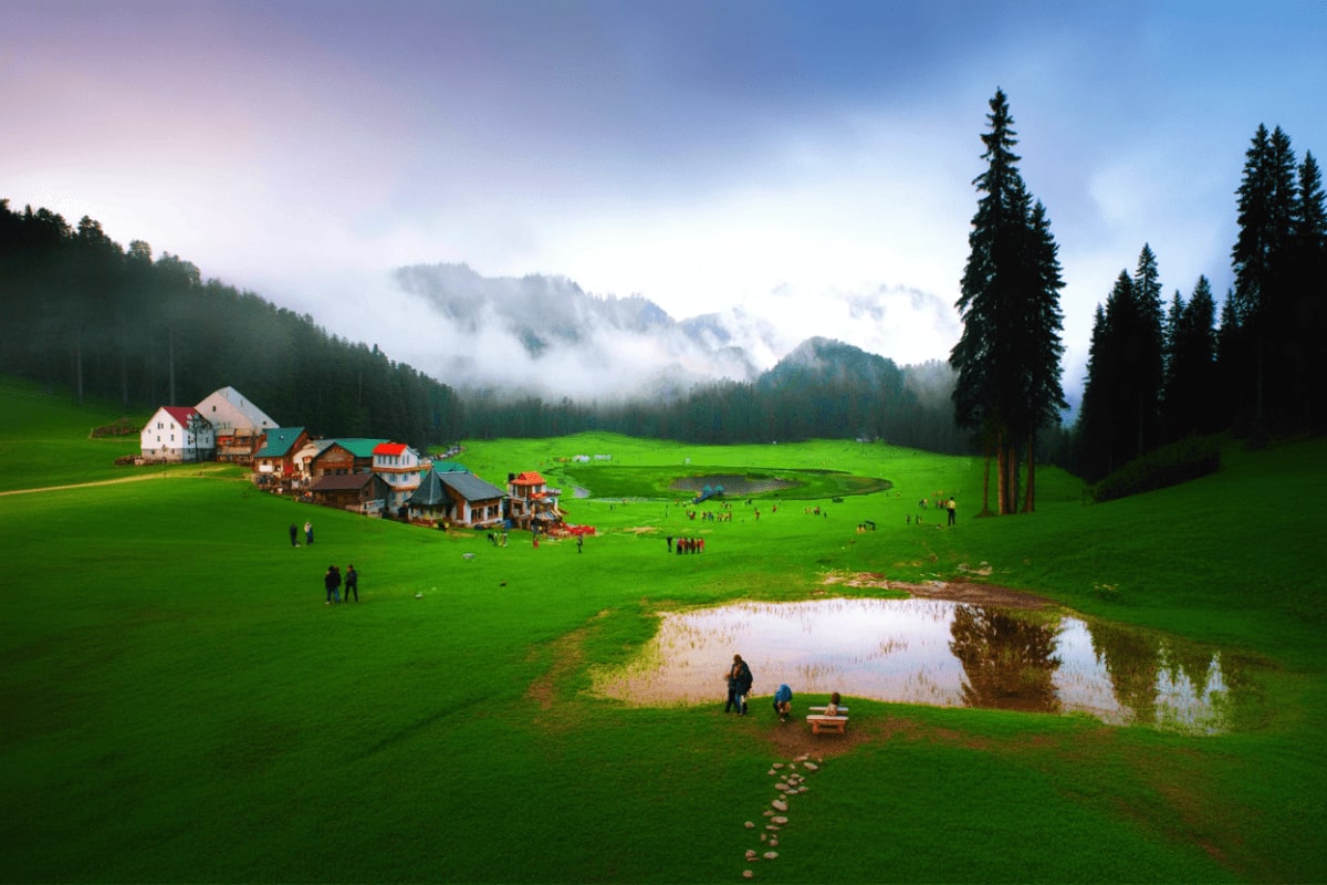 Best of Himachal with Amritsar from Dharamshala Dalhousie and Golden Temple Alpine meadow with traditional wooden houses, small pond, coniferous forest, and snow-capped mountain peaks in misty backgrou