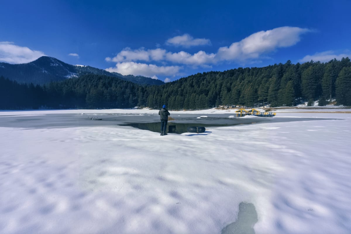 Person standing on frozen lake with pine forest-covered mountains, blue sky, and yellow boat houses visible in distance