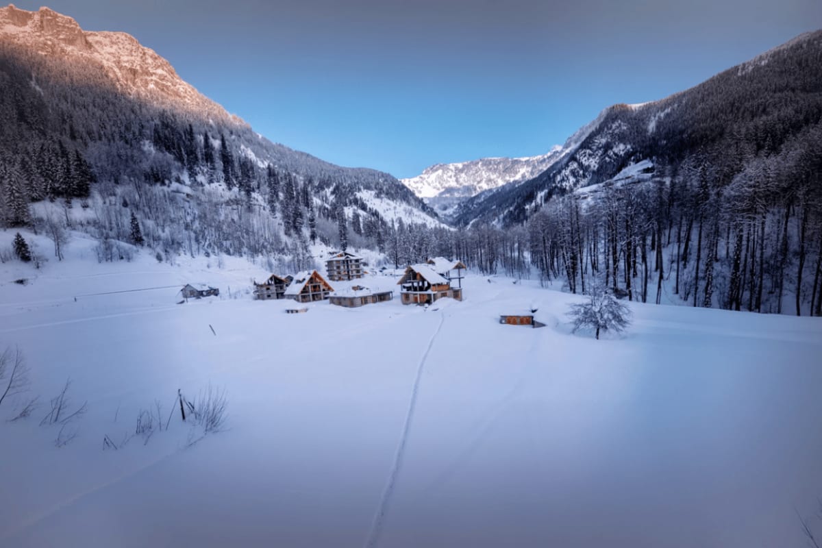 Snow-covered valley with wooden lodges nestled between forested mountains and snow-capped peaks in Himachal Pradesh