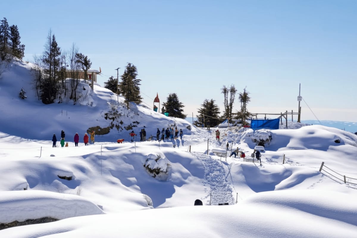 Snow-covered mountain valley with visitors exploring snowy landscape, temple with spire on hillside, evergreen trees, and cle