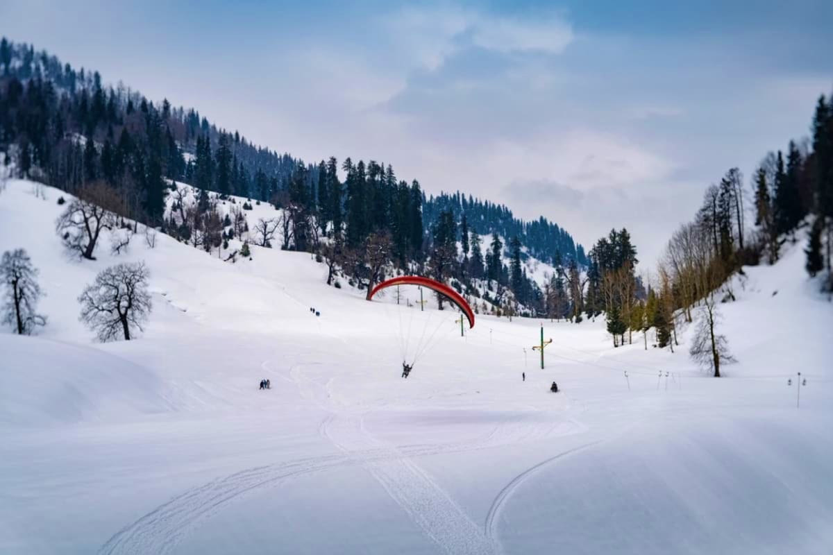 Snow Valleys And Hot Springs Tour In Manali And Manikaran Paraglider with red parachute flying over snow-covered valley with coniferous forests and snow-laden mountains in Himachal Pr
