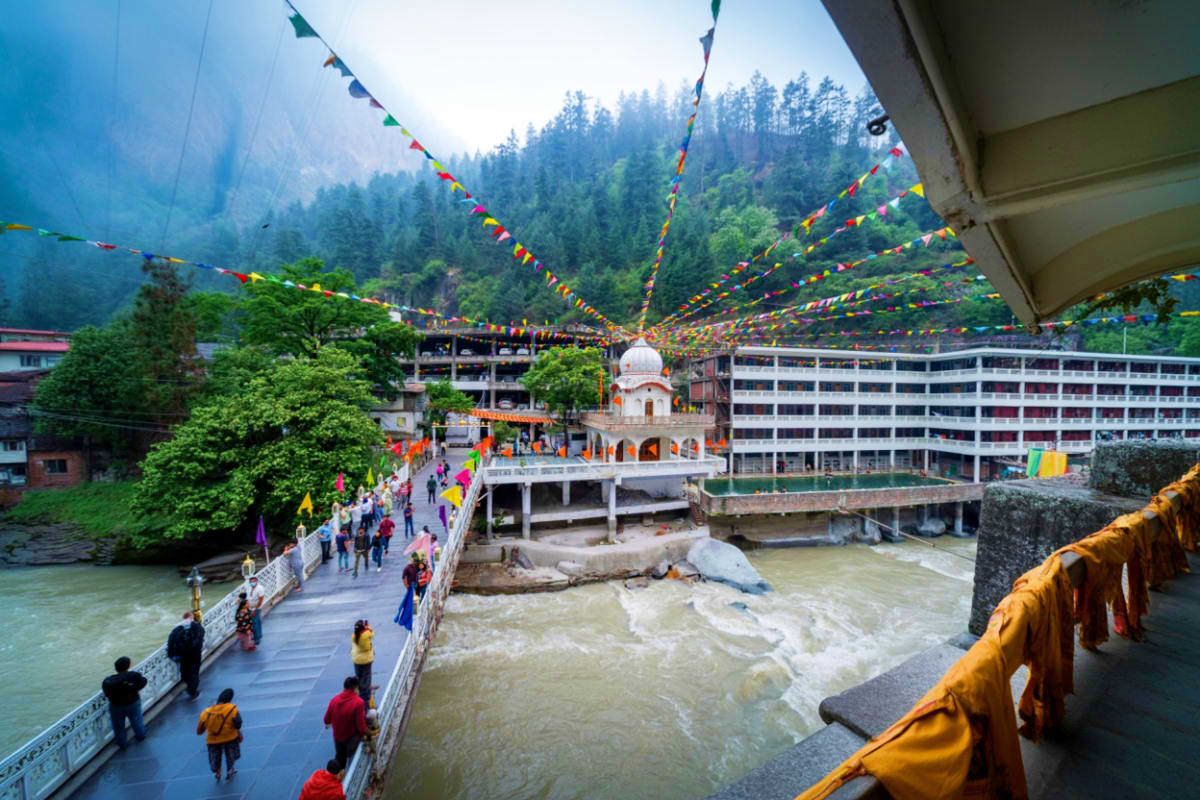 Snow Valleys And Hot Springs Tour In Manali And Manikaran Himalayan valley town with colorful prayer flag strings spanning overhead, white temple building, river, forested mountains,
