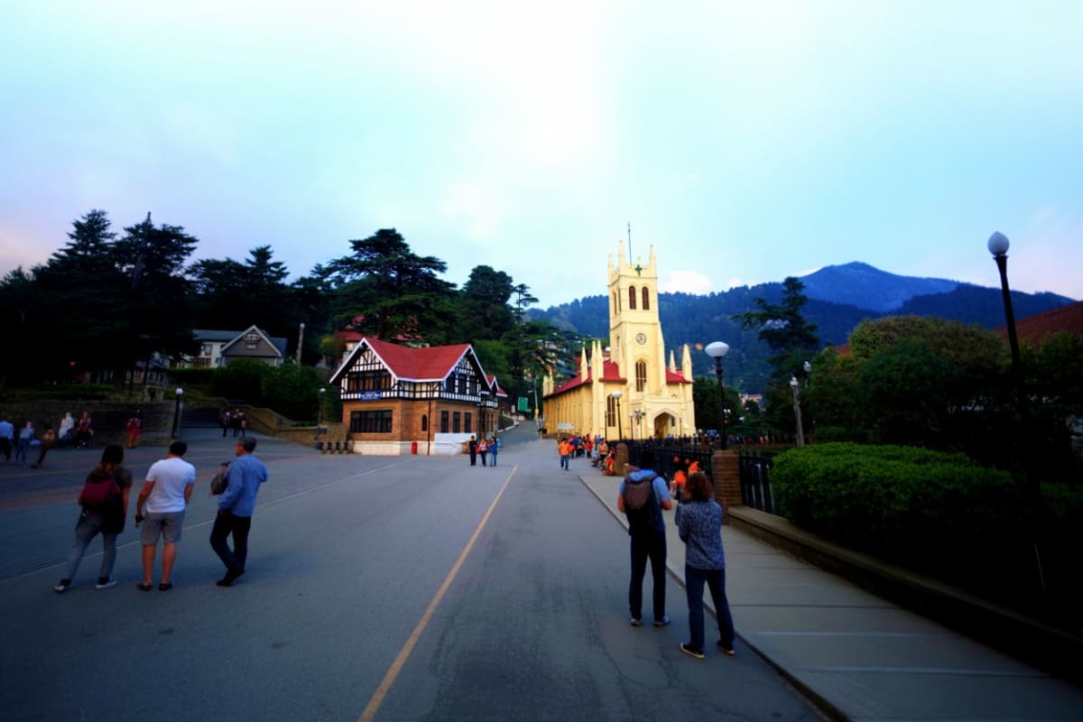 Snow Valleys And Hot Springs Tour In Manali And Manikaran Tourists walking on a street toward Christ Church, a yellow colonial-era church with mountains in the background in Shimla, I