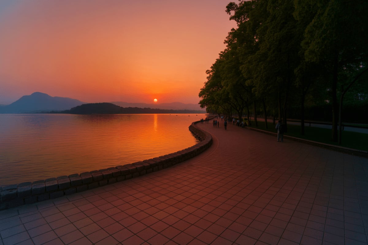Sunset over a waterfront promenade with curved seawall, tree-lined path, and mountains silhouetted against orange sky reflect